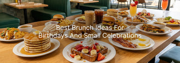 A long wooden table filled with brunch dishes including pancakes, waffles with fruit and cream, eggs, bacon, burgers, fries, tacos, iced coffee, and colorful drinks set for a birthday or small celebration.