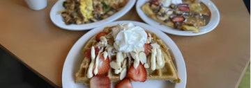 Alt text: Plate of waffles topped with sliced strawberries, whipped cream, and almond slices, with two other breakfast plates blurred in the background.