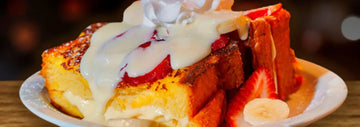 Thick brioche French toast topped with strawberries, whipped cream, and creamy sauce, served with banana slices on a white plate.