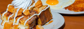 Thick French toast topped with whipped cream, white icing drizzle, caramel candy pieces, and caramel sauce, with a sunny-side up egg and hash browns in the background.