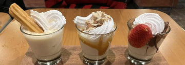 Three small dessert cups topped with whipped cream, a churro in the first, caramel and almond flakes in the second, and a strawberry with chocolate drizzle in the third.