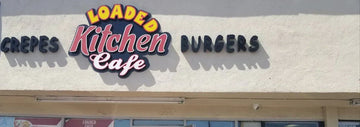 Exterior storefront sign reading “Loaded Kitchen Cafe” with “Crepes” on the left and “Burgers” on the right displayed on a beige building facade.